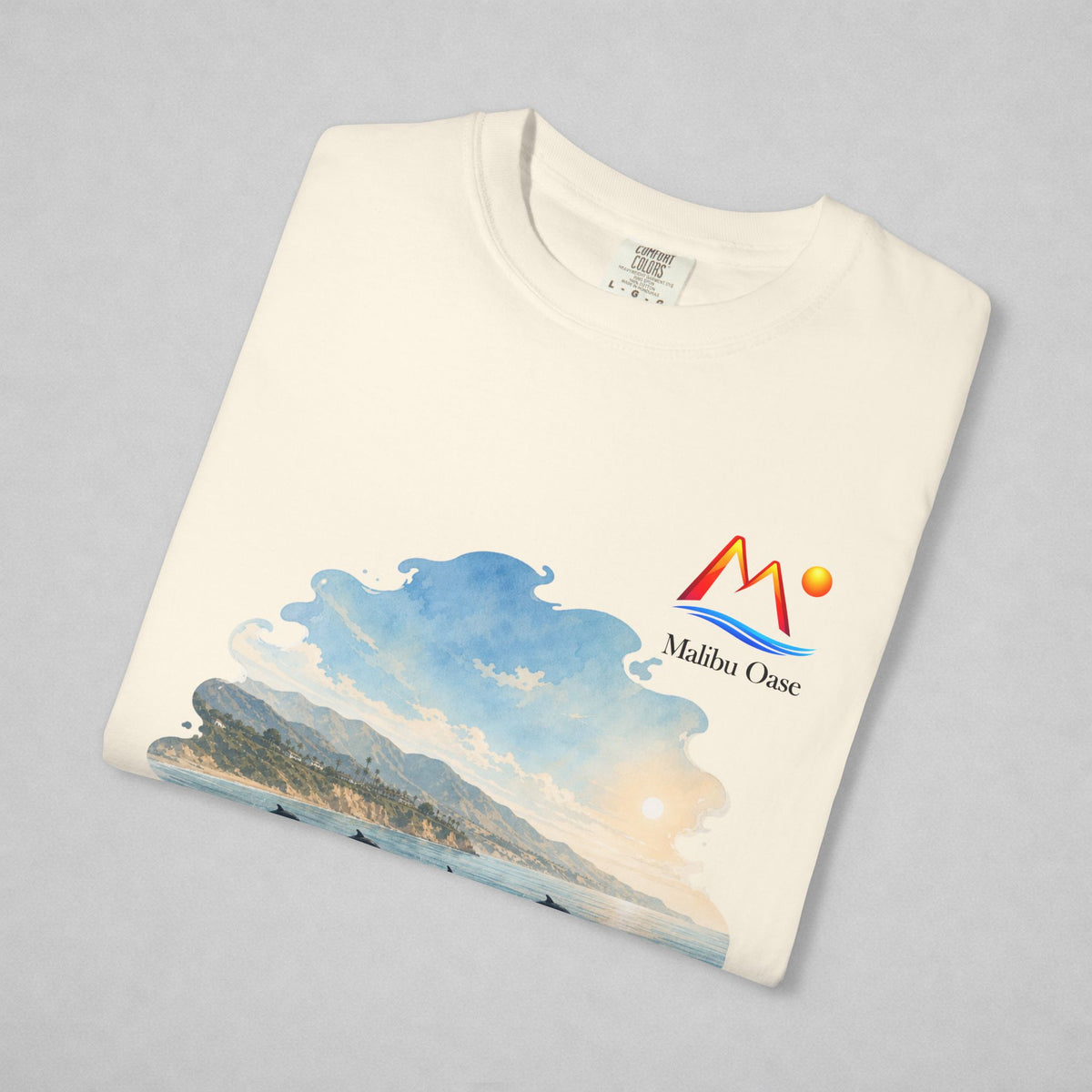 A folded Ivory t-shirt on a light gray background, featuring a colorful graphic print of a coastal landscape with mountains, waves, and a sun, alongside the 'Malibu Oase' logo.