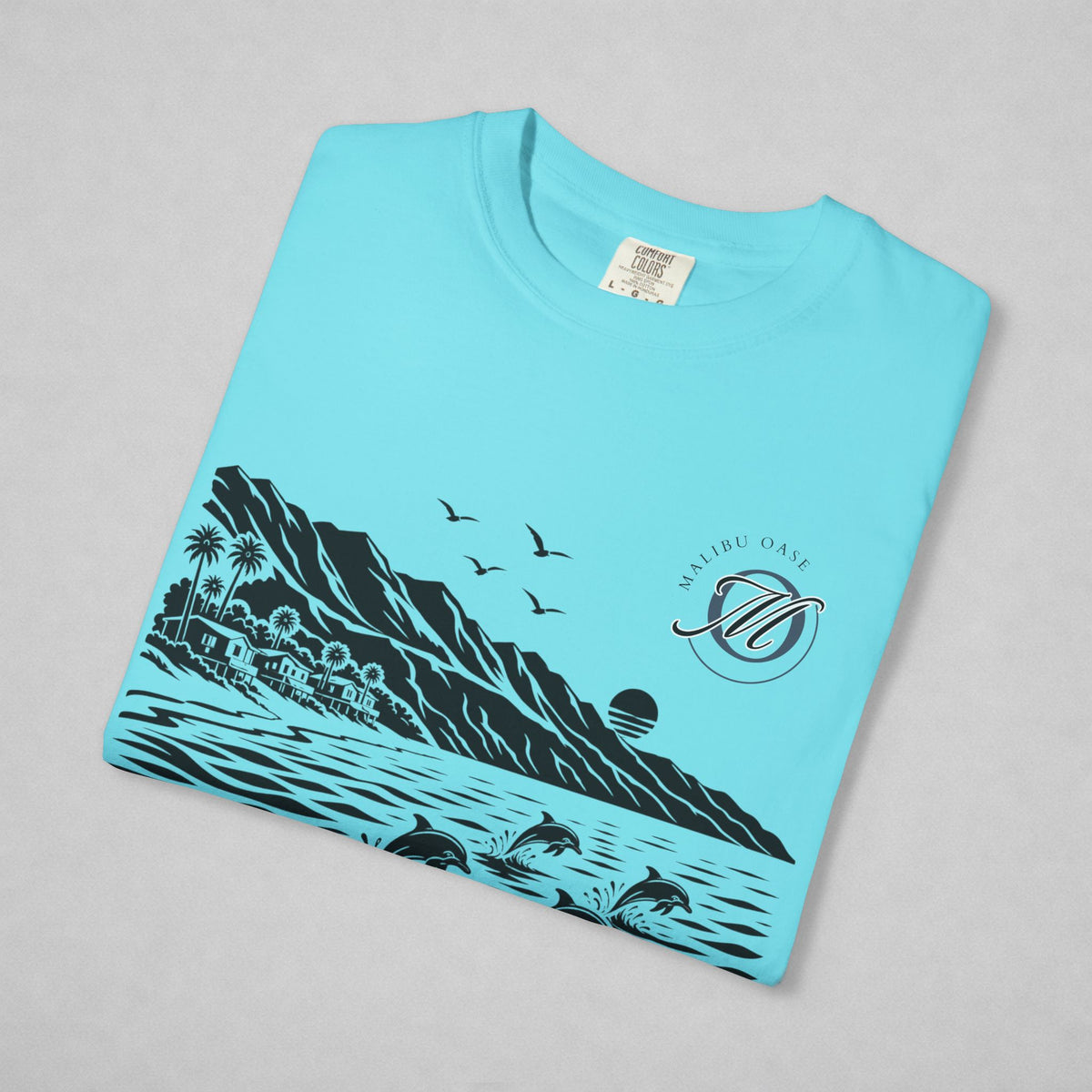 A folded light blue t-shirt with a black graphic print of a coastal landscape, including mountains, palm trees, birds, dolphins, a sun and a "Malibu Oase" monogram.