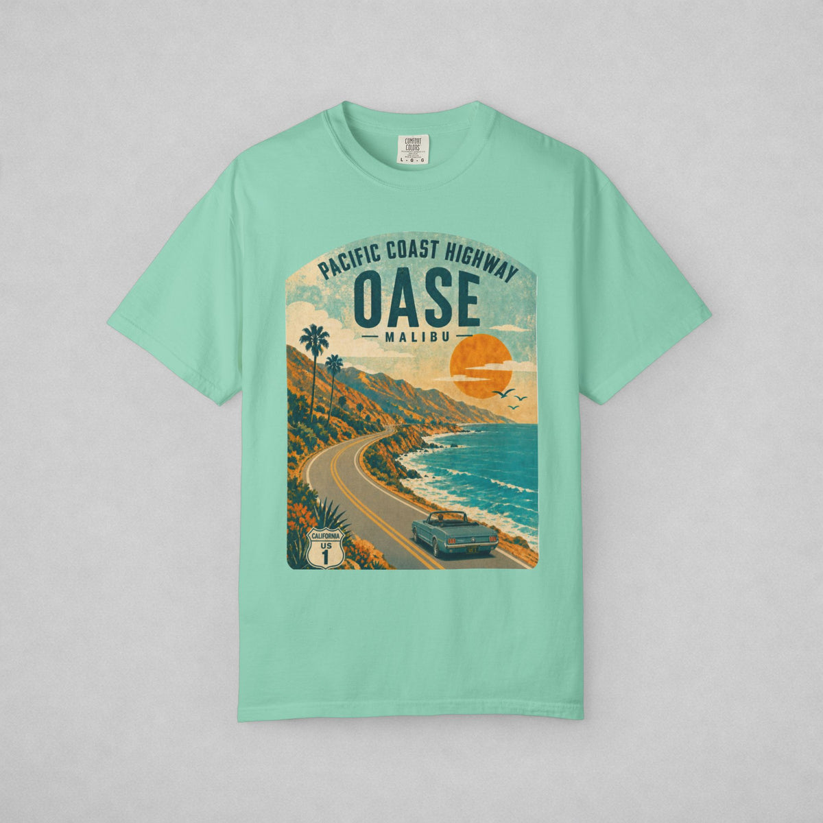 A mint green short-sleeve t-shirt with a vintage-style graphic print of the Pacific Coast Highway Malibu. The graphic features a retro sunset, palm trees, a winding coastal road, and a car, set against a light gray background.