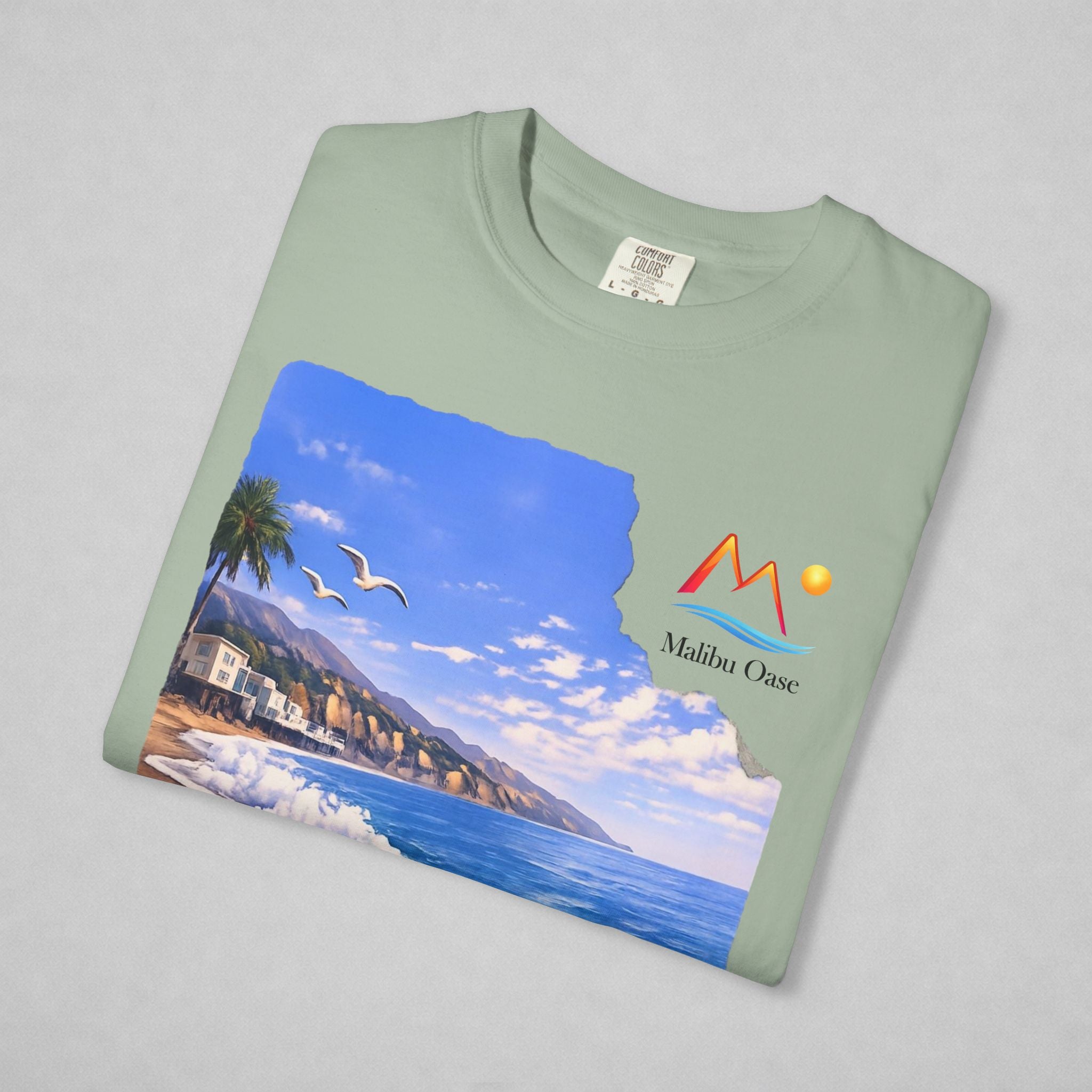 A folded sage t-shirt on a neutral gray background, featuring a printed graphic of a coastal landscape with a mountain logo that says 'Malibu Oase.'