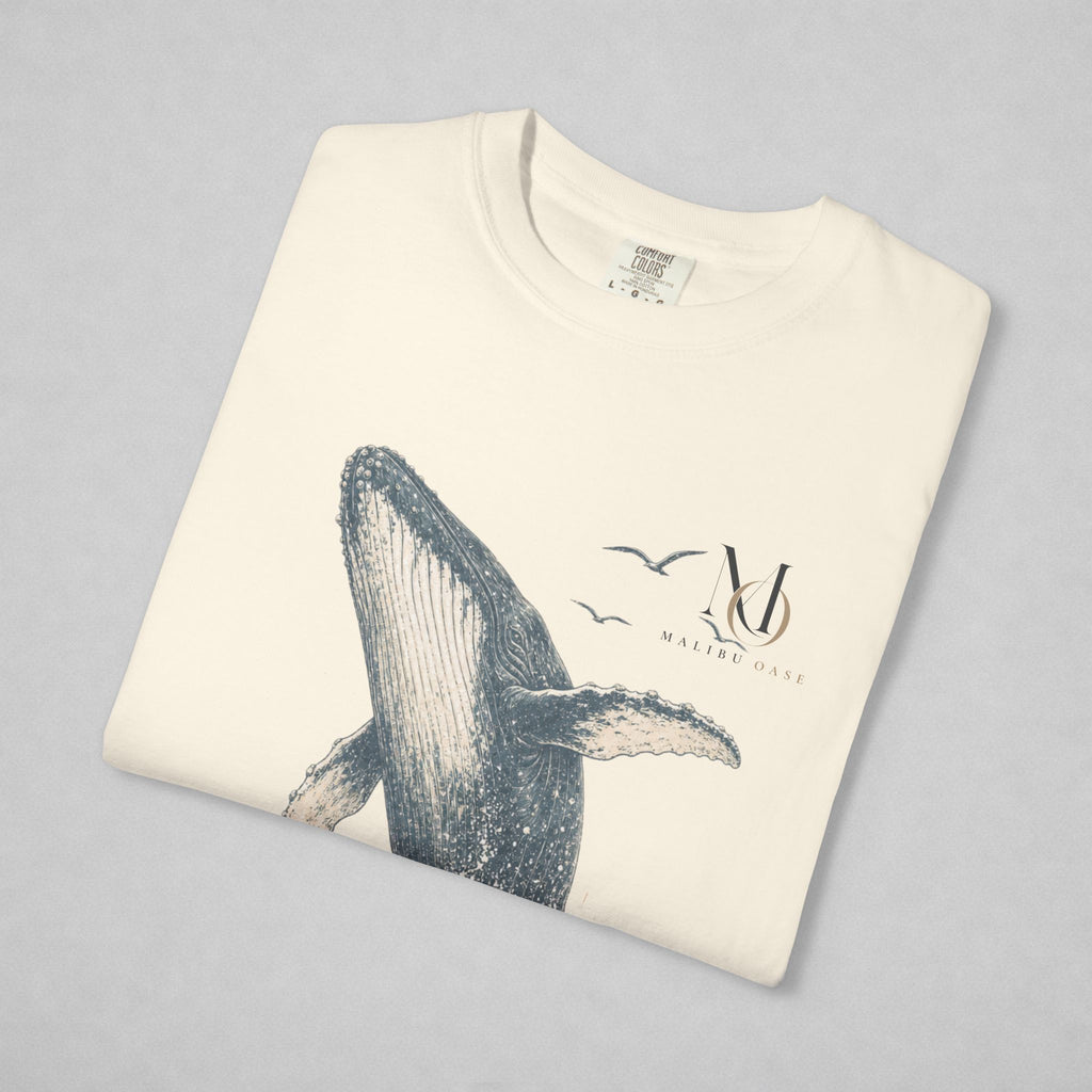 A folded cream-colored t-shirt with a graphic print of a humpback whale, birds, and a stylized 'Malibu Oase' monogram on the chest, displayed against a neutral gray background.