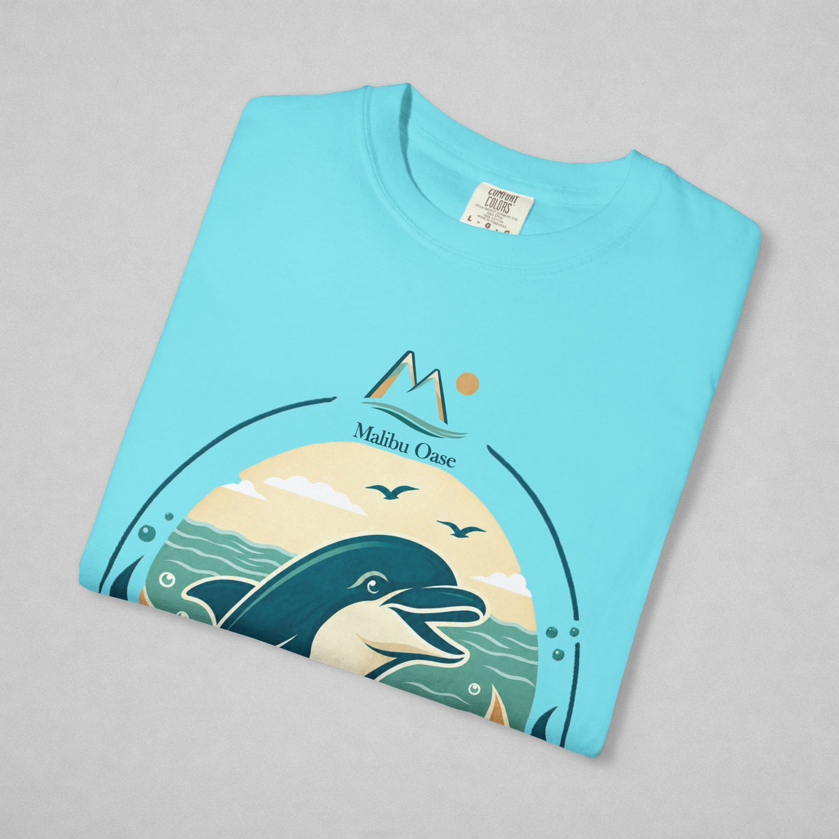 A folded light blue t-shirt on a neutral gray background, featuring a graphic print of a dolphin in a Malibu Ocean scene with mountains and birds.