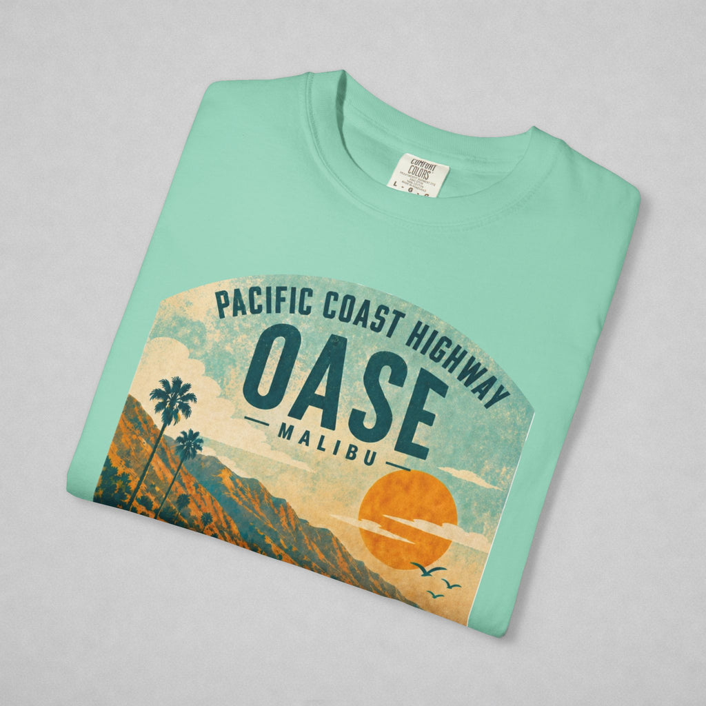 A folded mint green t-shirt with a vintage-style graphic of the Pacific Coast Highway Oase Malibu, featuring a sunset, palm trees, and mountains, displayed on a light gray background.