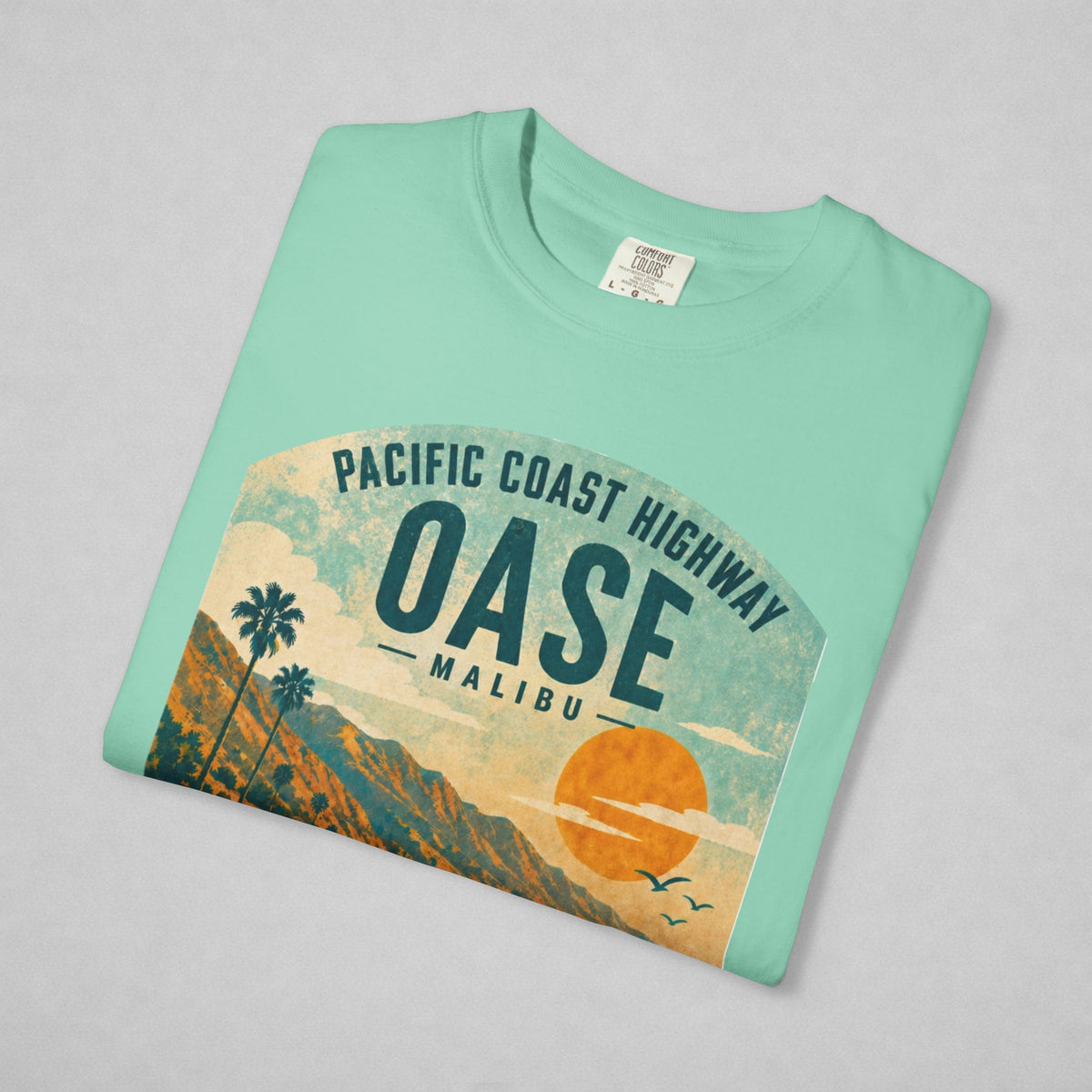 A folded mint green t-shirt with a vintage-style graphic of the Pacific Coast Highway Oase Malibu, featuring a sunset, palm trees, and mountains, displayed on a light gray background.