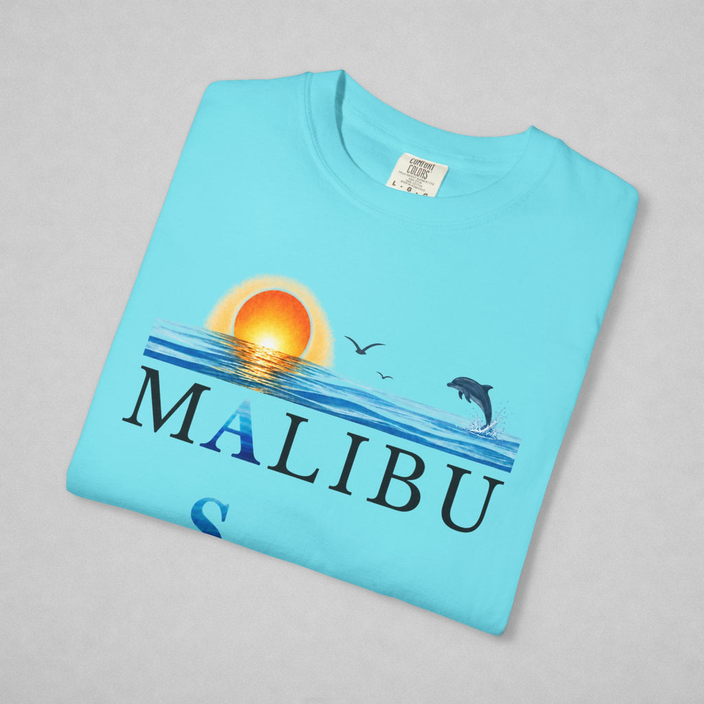A folded light blue t-shirt on a neutral gray background, featuring a graphic print of a sunset over ocean waves with a jumping dolphin, seagulls, and the word 'MALIBU' printed across the front.