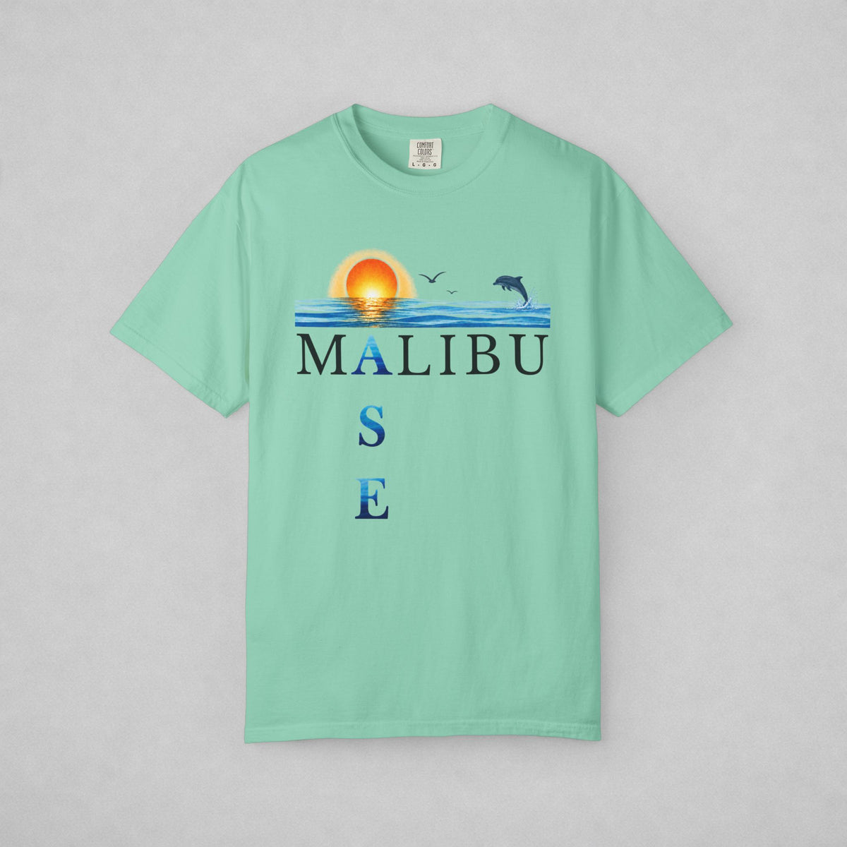 A mint green short-sleeve t-shirt with a 'MALIBU OASE' graphic on the chest, featuring a sunset, ocean waves, birds, and dolphins, displayed flat against a light gray background.