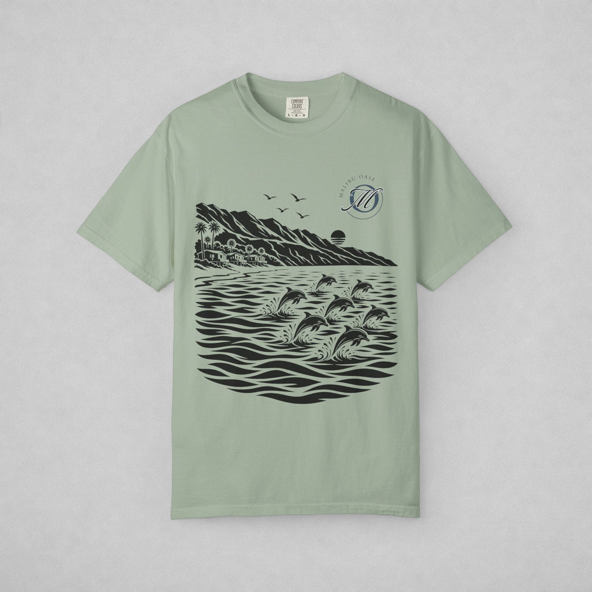 A sage green short-sleeve t-shirt with a black graphic print of dolphins jumping in water, mountains, birds, and a sun, displayed flat against a light gray background.