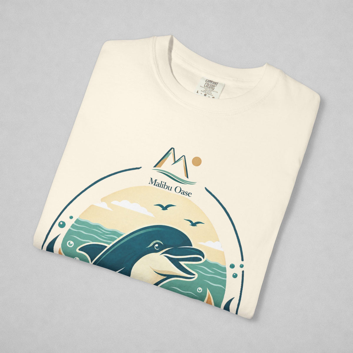 A folded cream-colored t-shirt with a graphic print of a dolphin, waves, mountains, and the text 'Malibu Oase' on a light gray background.