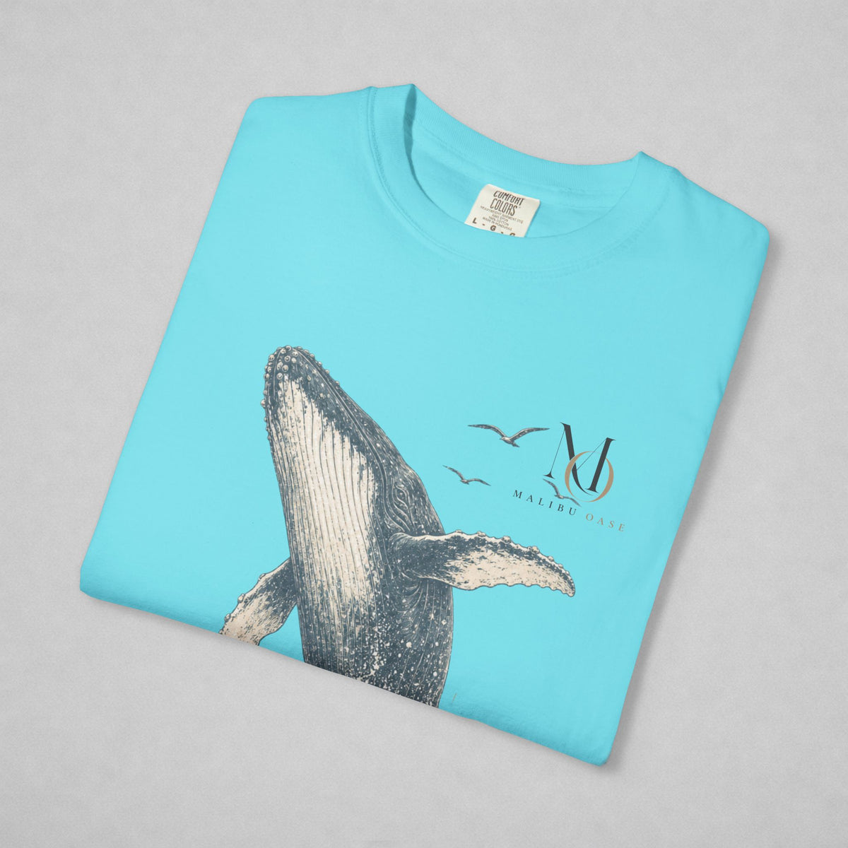 A folded light blue t-shirt with a graphic print of a whale, birds, and a stylized 'M' logo with 'MALTESE MARINE' underneath, displayed on a neutral gray background.