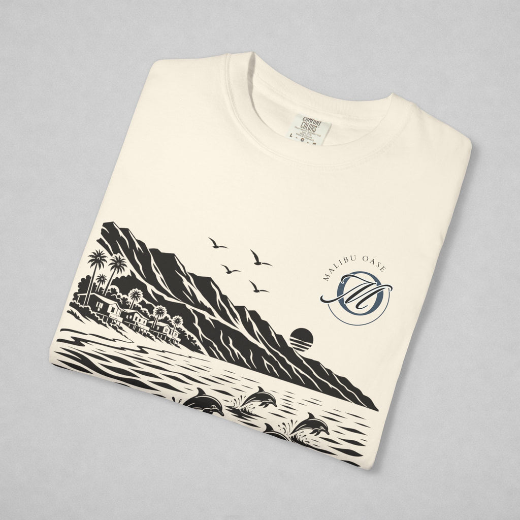 An off-white crewneck sweatshirt folded neatly on a light gray background, featuring a black and navy blue graphic print of a coastal landscape with mountains, dolphins, birds, and a sun, alongside a circular monogram that reads 'Malibu Oase'.