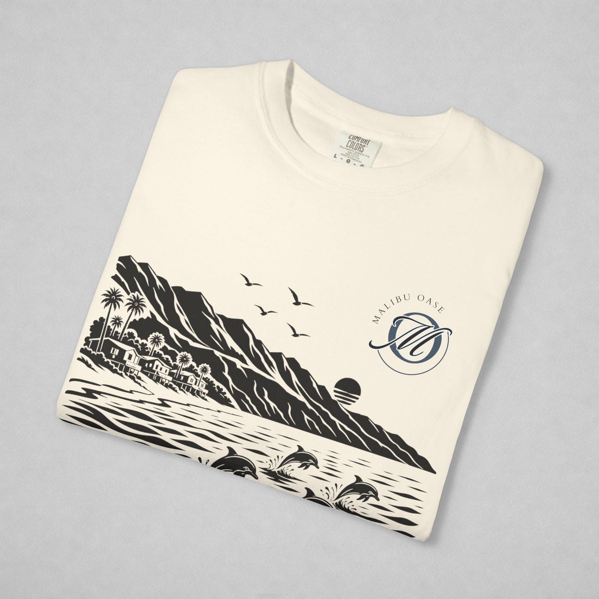An off-white crewneck sweatshirt folded neatly on a light gray background, featuring a black and navy blue graphic print of a coastal landscape with mountains, dolphins, birds, and a sun, alongside a circular monogram that reads 'Malibu Oase'.