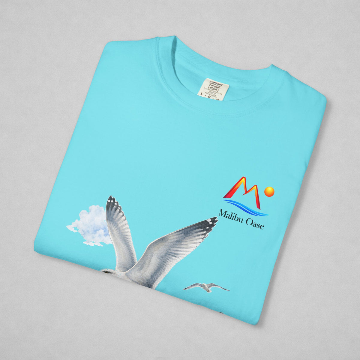A folded light blue t-shirt on a neutral gray background, featuring a graphic print of a flying seagull and a 'Malibu Oase' logo on the front.