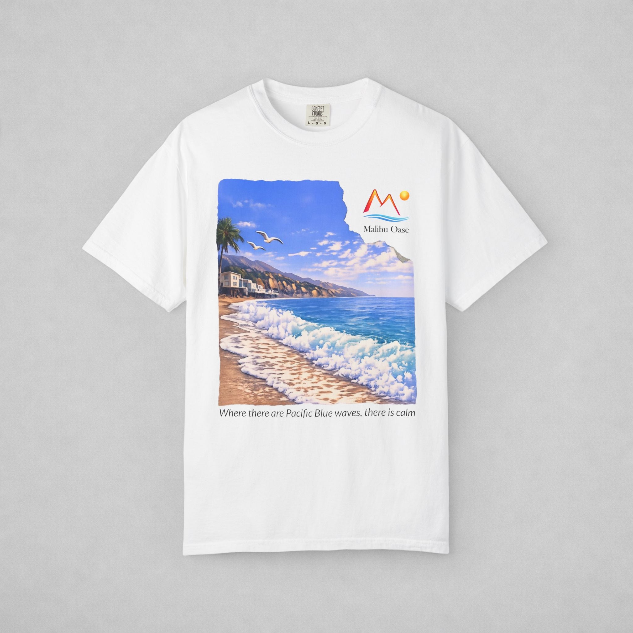 A white short-sleeve crewneck t-shirt with a large graphic print of a beach scene on the front. The graphic features a blue sky, ocean waves, a sandy beach, and a palm tree, with the logo 'Malibu Oase' and under the print there are the words 'Where there are Pacific Blue waves, there is calm'.
