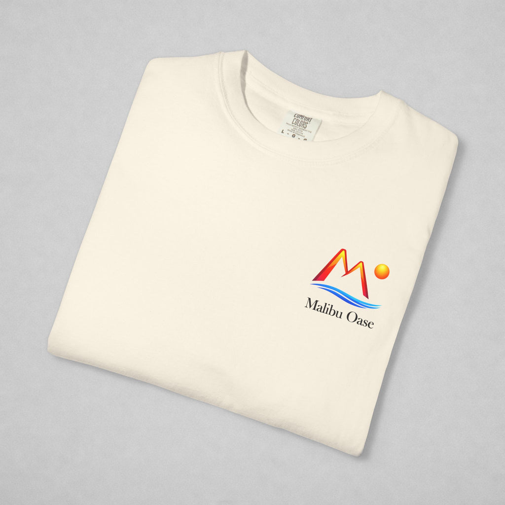 A folded off-white crewneck t-shirt on a light gray background, featuring a colorful 'Malibu Oasis' logo on the left chest with red, orange, and blue graphic elements.