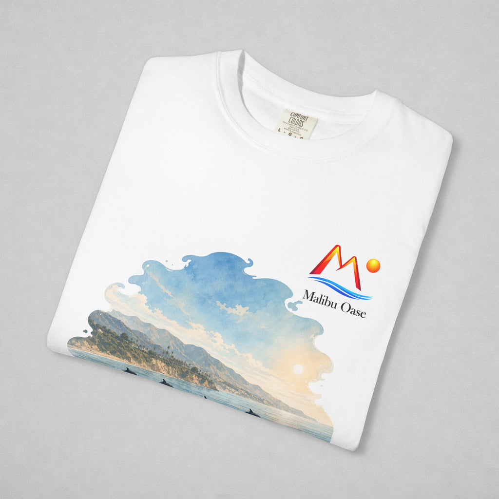 A folded white t-shirt on a light gray background, featuring a colorful graphic print of a coastal landscape with mountains, waves, and a sun, alongside a red and blue 'Malibu Oase' logo.