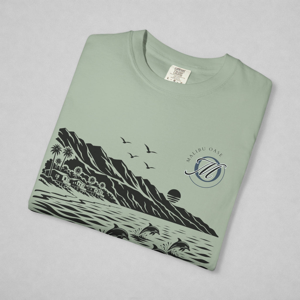 A folded sage green t-shirt with a black and gray graphic print of a tropical beach scene, featuring mountains, palm trees, dolphins, birds, and a sun. The monogram 'Malibu Oase' is printed on the chest.