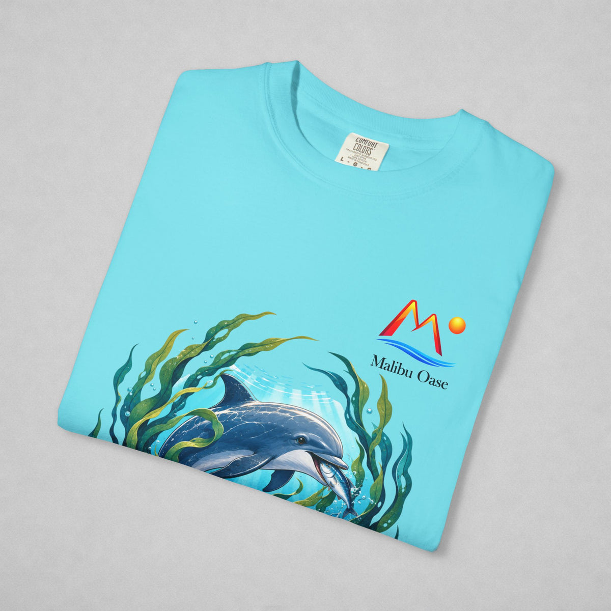 A folded light blue t-shirt on a neutral gray background, featuring a graphic print of a dolphin swimming among green seaweed, with a 'Malibu Oase' logo printed on the chest.