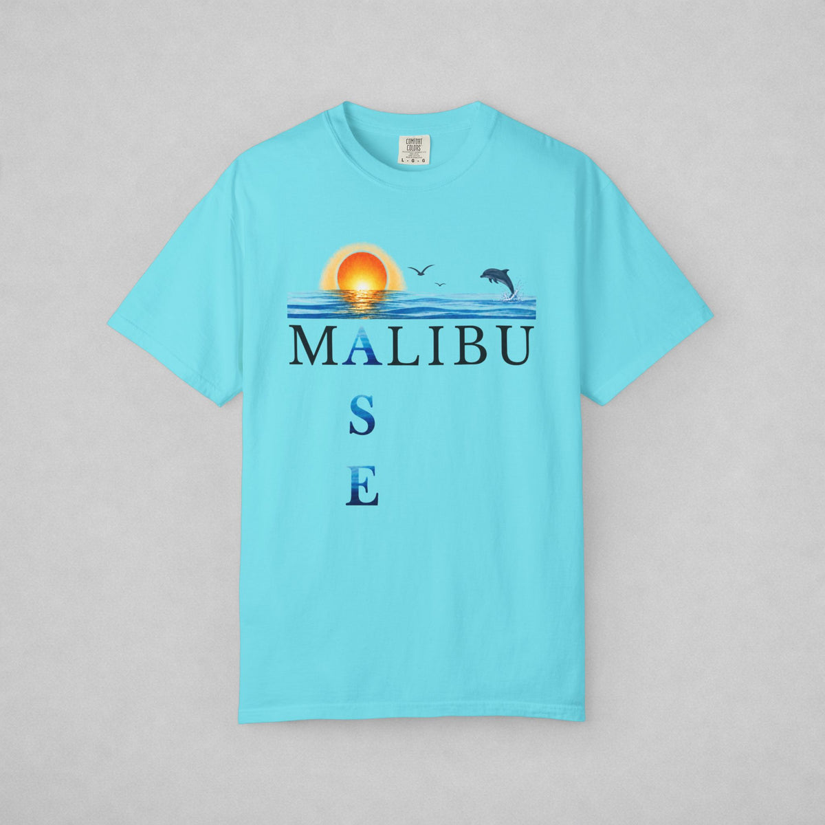 A light blue short-sleeve t-shirt with a graphic on the chest depicting a sunset over water, two birds, a dolphin, and the text 'MALIBU OASE'. The shirt is displayed flat against a neutral gray background.