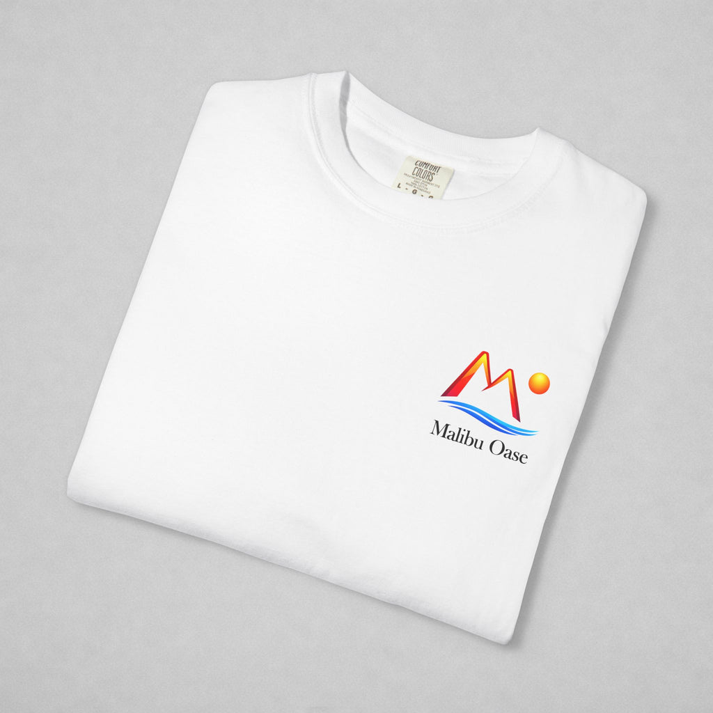 A folded white crewneck t-shirt on a light gray background, featuring a colorful 'Malibu Oase' graphic on the left chest with red, orange, and blue mountain and wave elements.