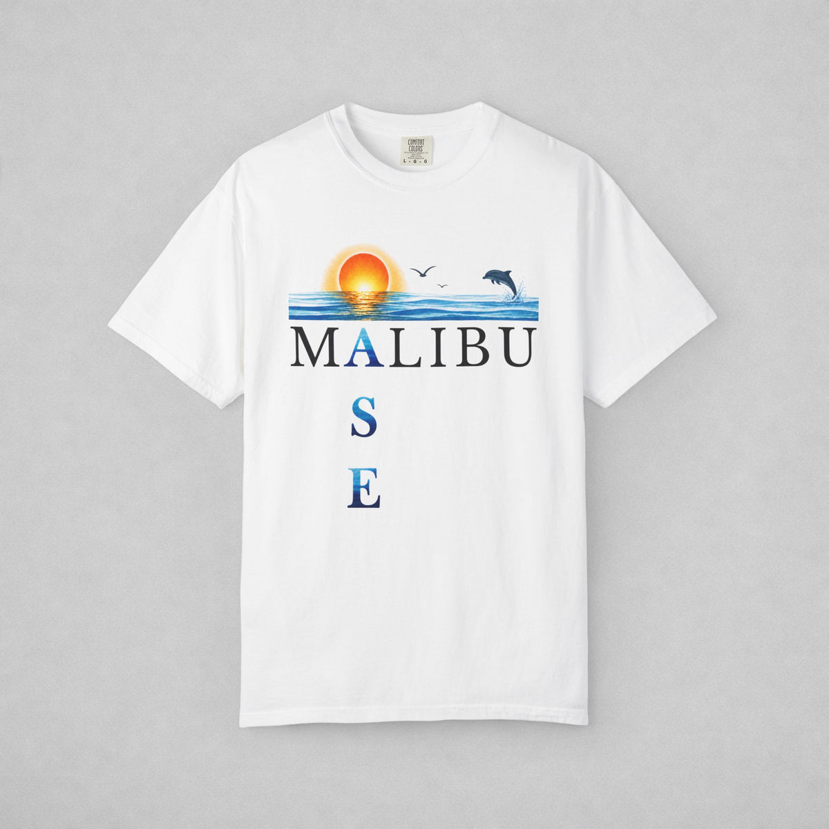 A white short-sleeve crewneck t-shirt featuring a graphic print of a sunset over blue water with a dolphin, birds, and the text 'MALIBU OASE', displayed on a neutral gray background.