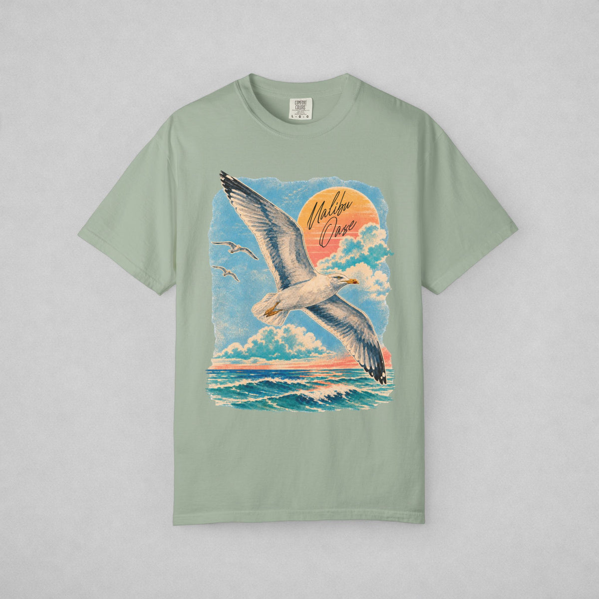 A sage green short-sleeve t-shirt with a large graphic print of a seagull flying over the ocean under a blue sky with a pink and orange sunset. The words 'Malibu Oase' are written in script on the graphic.