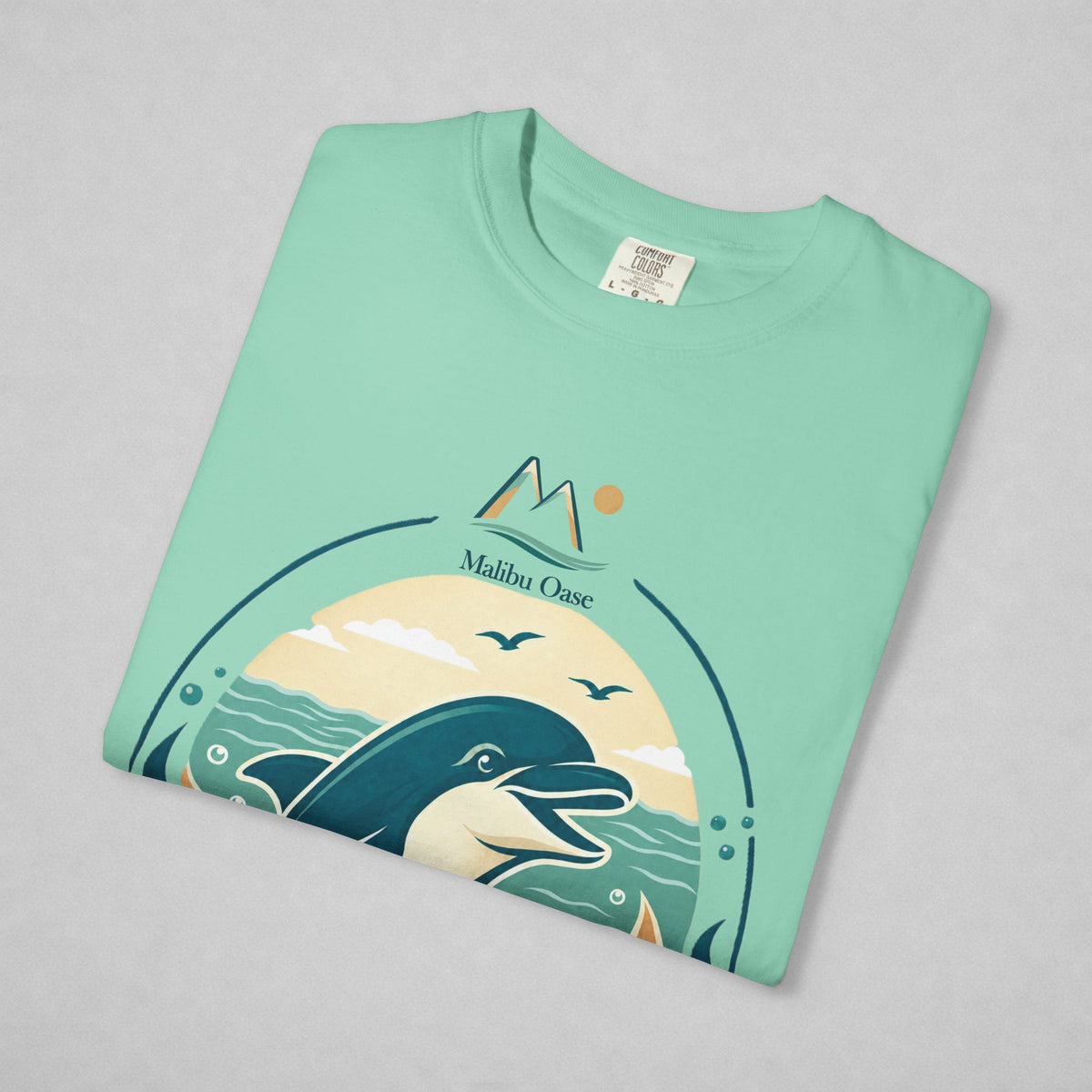 A folded mint green t-shirt with a 'Malibu Oase' graphic print featuring a dolphin, mountains, and a sunset, displayed on a neutral gray background.