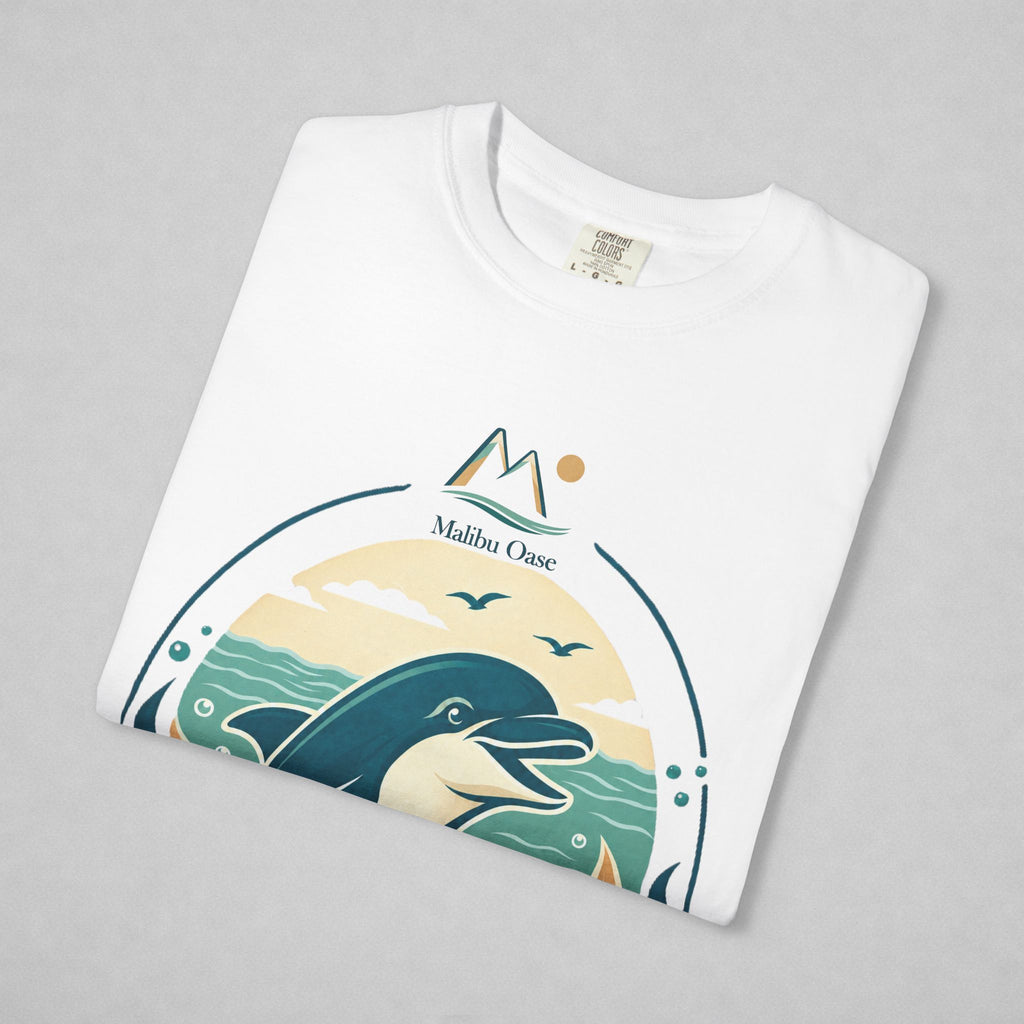 A folded white crewneck t-shirt with a graphic print of a dolphin, mountains, and the text 'Malibu Oase' on a light gray background.