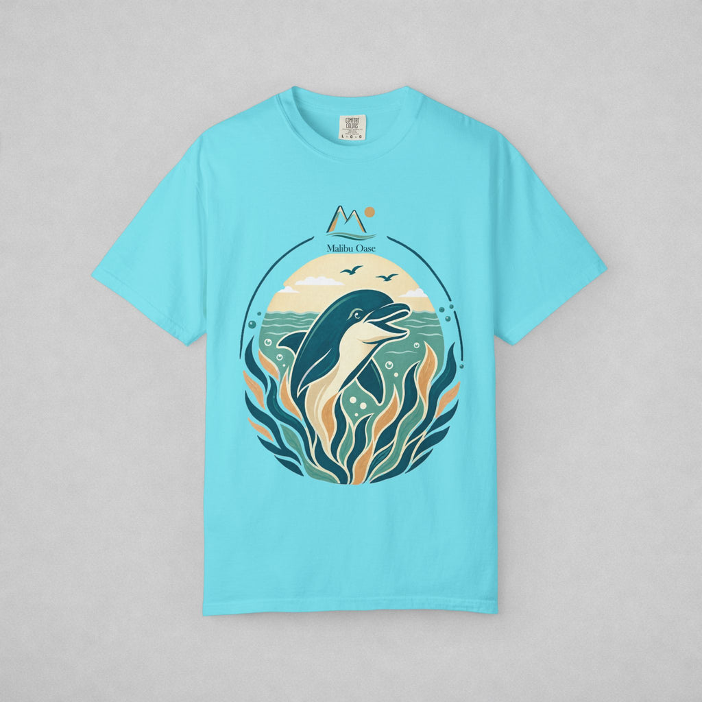 A light blue short-sleeve t-shirt with a circular graphic print of a dolphin swimming among stylized waves and seaweed, featuring the text 'Malibu Oase' and a mountain icon above. The t-shirt is displayed flat against a neutral gray background.