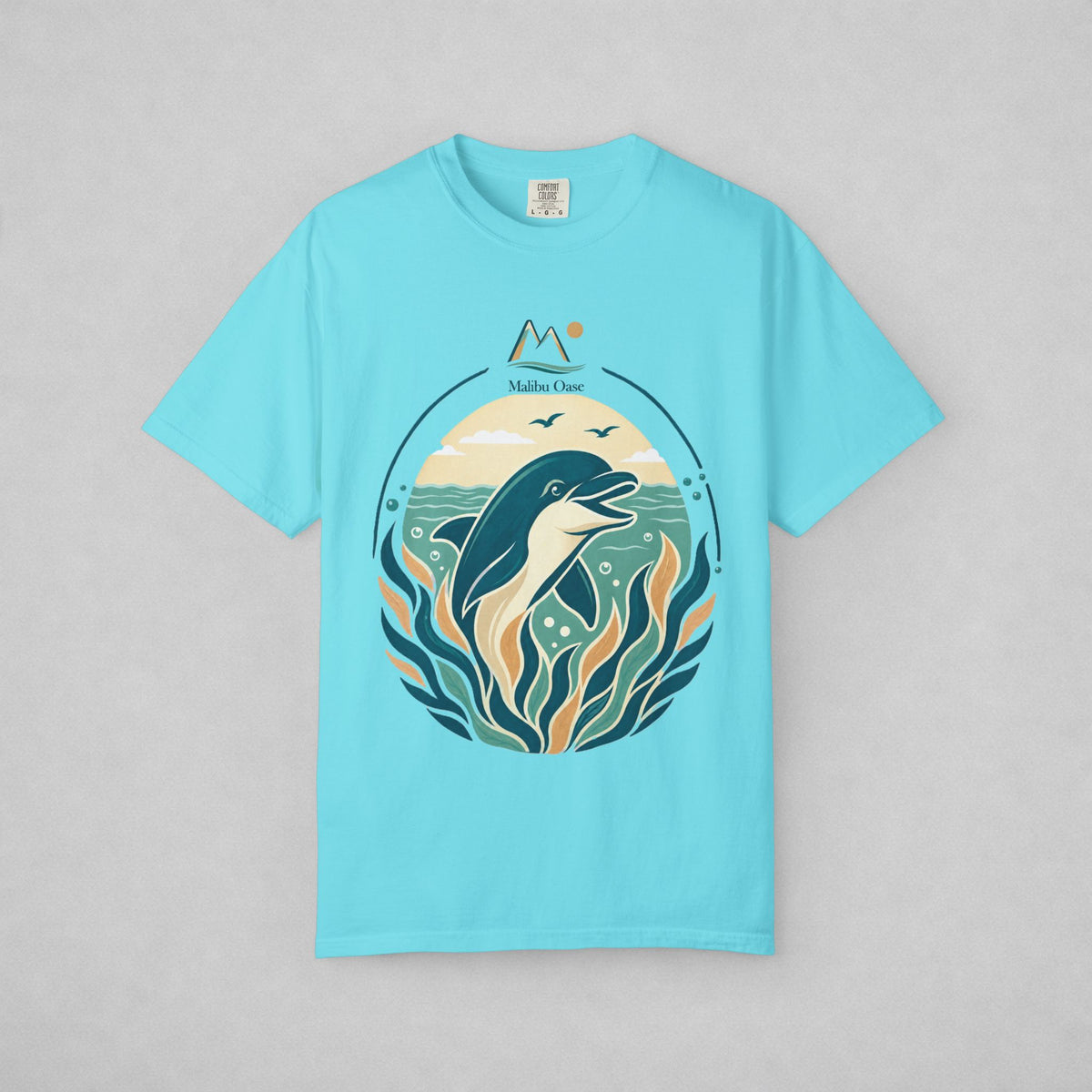 A light blue short-sleeve t-shirt with a circular graphic print of a dolphin swimming among stylized waves and seaweed, featuring the text 'Malibu Oase' and a mountain icon above. The t-shirt is displayed flat against a neutral gray background.