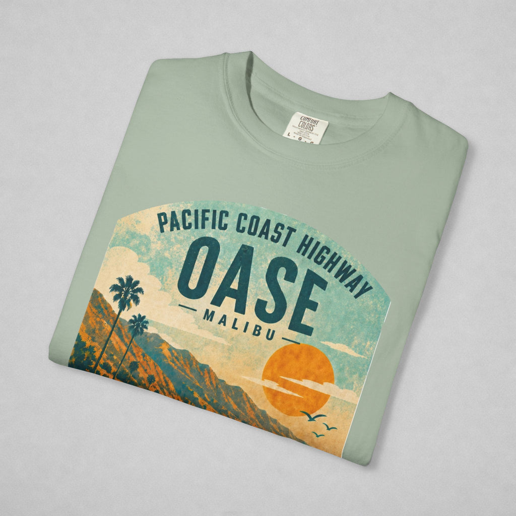 A folded sage green t-shirt with a retro graphic of the Pacific Coast Highway Oase Malibu, displayed on a neutral gray background.