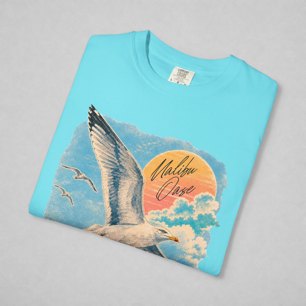 A folded light blue t-shirt on a neutral gray background, featuring a graphic print of a flying seagull, smaller birds, clouds, and a sunset circle with the text 'Malibu Oase'.