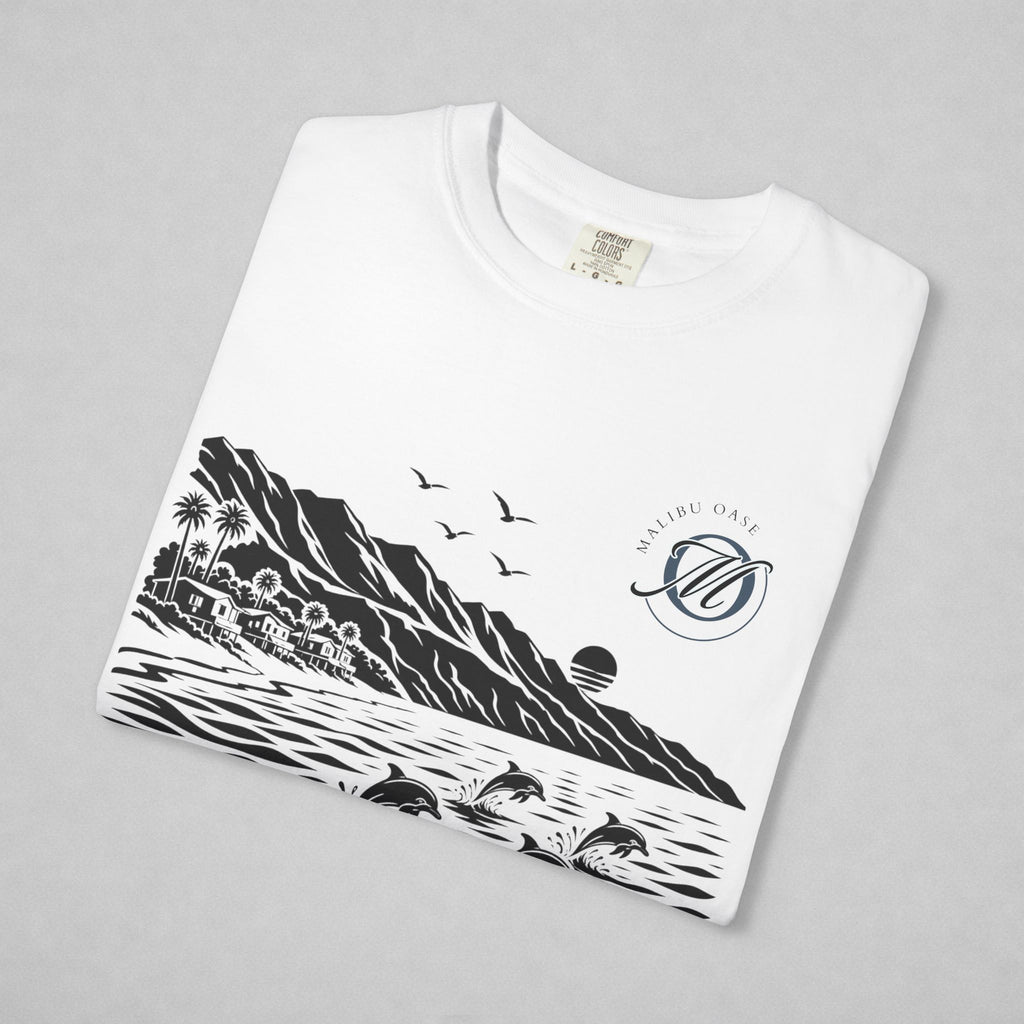 A folded white crewneck t-shirt with a black graphic print of a coastal landscape, including mountains, palm trees, a sunset, dolphins, birds and a "Malibu Oase" monogram.