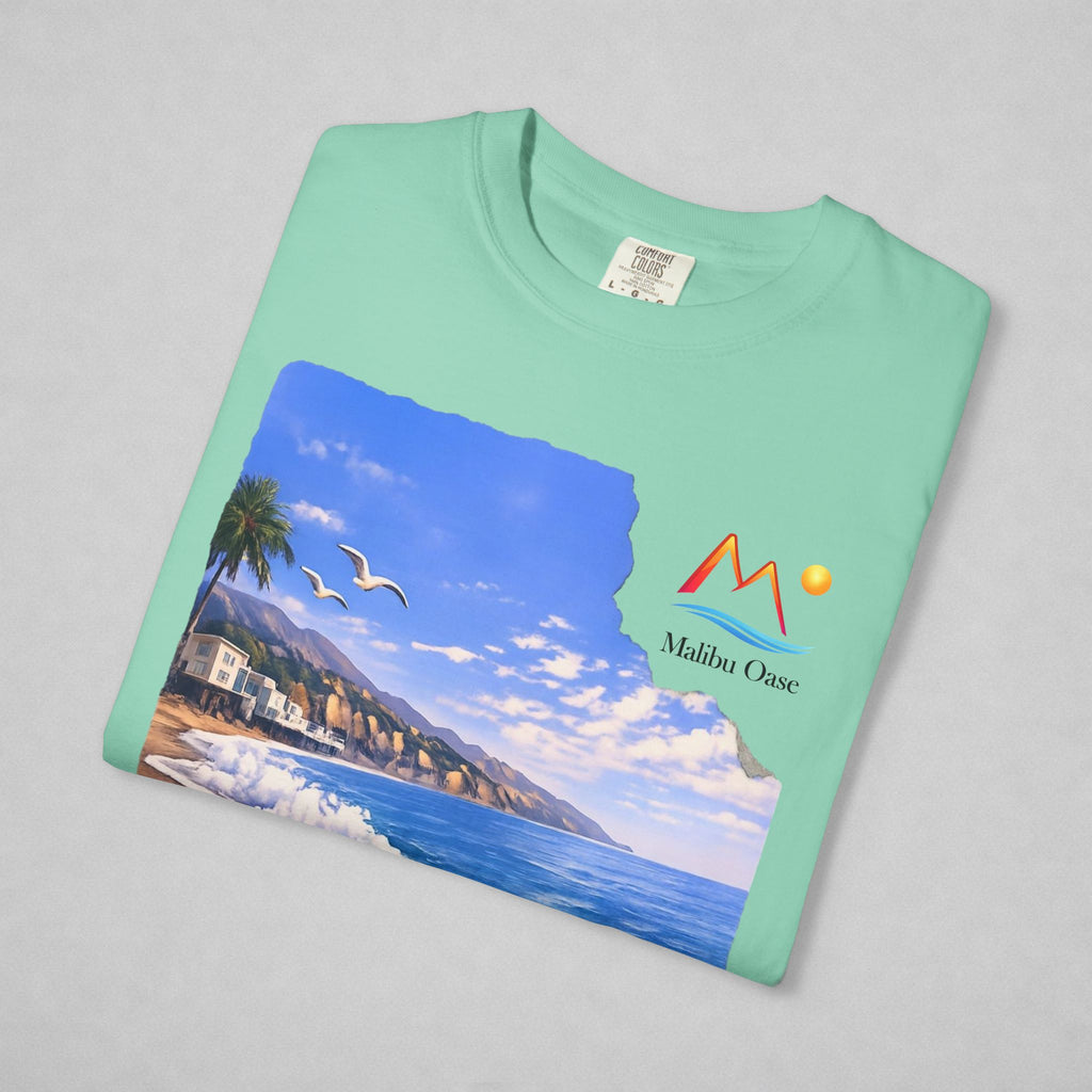 A folded mint green t-shirt on a neutral gray background, featuring a printed graphic of a coastal landscape with a mountain logo that says 'Malibu Oase.'