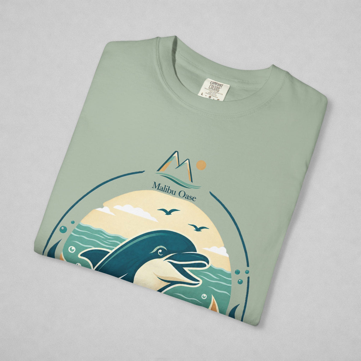A folded sage green t-shirt with a 'Malibu Oase' graphic print featuring a dolphin, mountains, and seagulls, displayed on a neutral gray background.