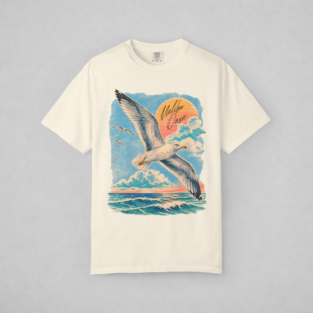 An off-white short-sleeve crewneck t-shirt featuring a large graphic print of a seagull flying over ocean waves under a blue sky with a pink and orange sunset. The words 'Malibu Oase' are written in cursive script on the graphic.