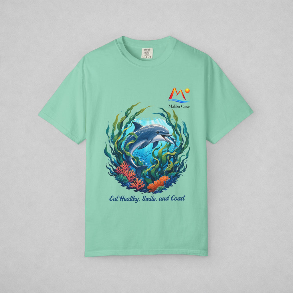 A mint green short-sleeve t-shirt with a colorful graphic of a dolphin swimming underwater with coral and seaweed, featuring the text 'Eat Healthy, Smile, and Coast' and a 'Malibu Oase' logo on the chest, displayed on a neutral gray background.