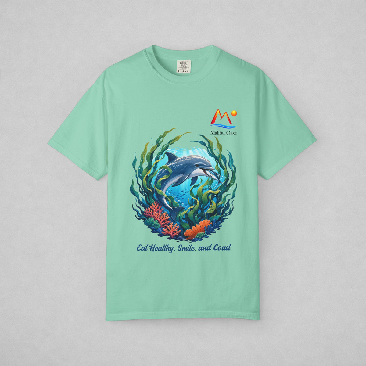 A mint green short-sleeve t-shirt with a colorful graphic of a dolphin swimming underwater with coral and seaweed, featuring the text 'Eat Healthy, Smile, and Coast' and a 'Malibu Oase' logo on the chest, displayed on a neutral gray background.