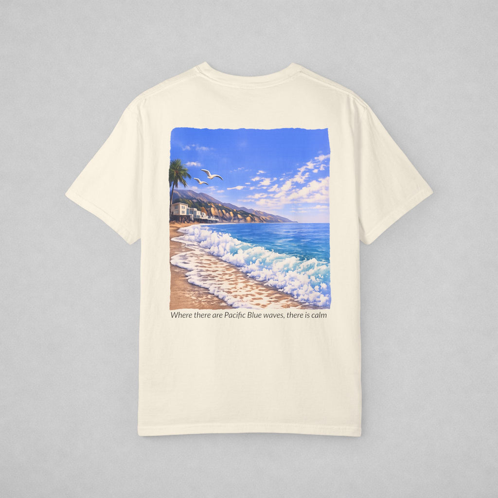 An off-white short-sleeve t-shirt with a large square graphic print of a beach scene featuring blue ocean waves, a sandy shore, and a blue sky. Below the graphic, the text reads 'Where there are Pacific Blue waves, there is calm'.