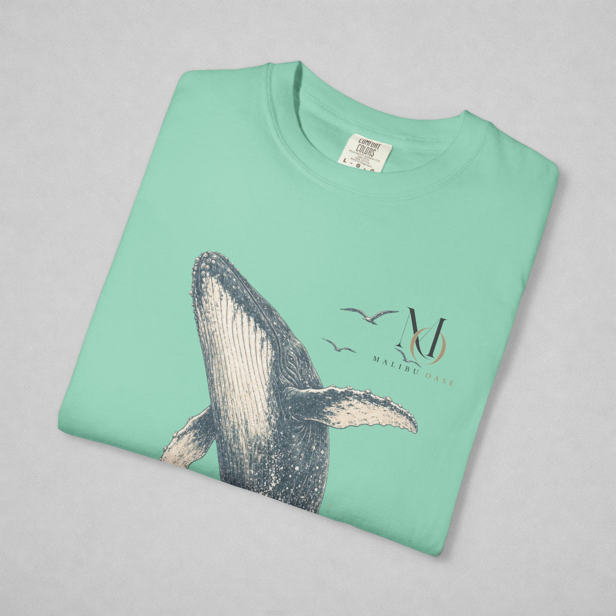 A folded mint green t-shirt on a light gray background, featuring a graphic print of a humpback whale, two birds, and a stylized 'Malibu Oase' monogram.