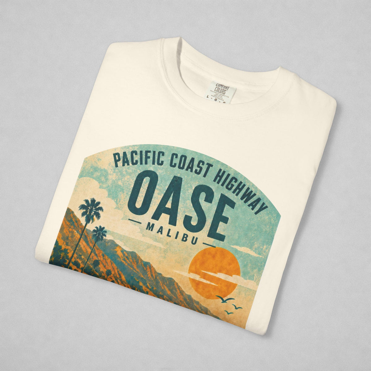 A folded cream-colored t-shirt with a retro graphic of the Pacific Coast Highway Oase Malibu, displayed on a neutral gray background.