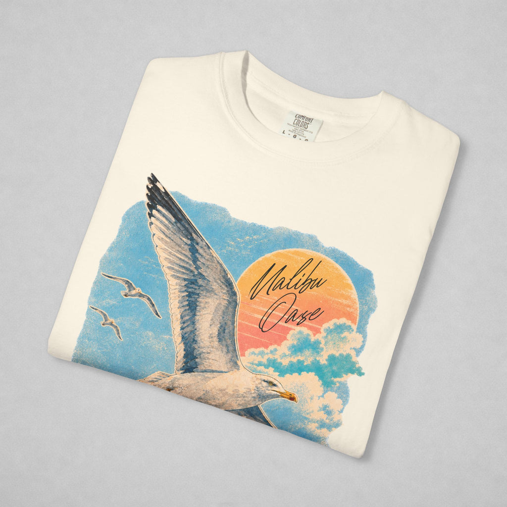 A folded off-white crewneck t-shirt with a colorful graphic print of a seagull flying over clouds and a sunset, featuring the text 'Malibu Oase'.
