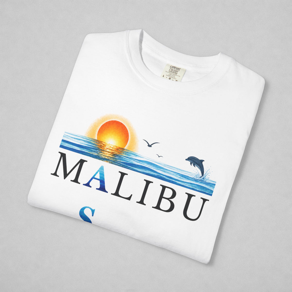 A folded white t-shirt with a colorful graphic print featuring a sunset, ocean waves, a jumping dolphin, seagulls, and the word 'MALIBU' in large letters, displayed on a neutral gray background.