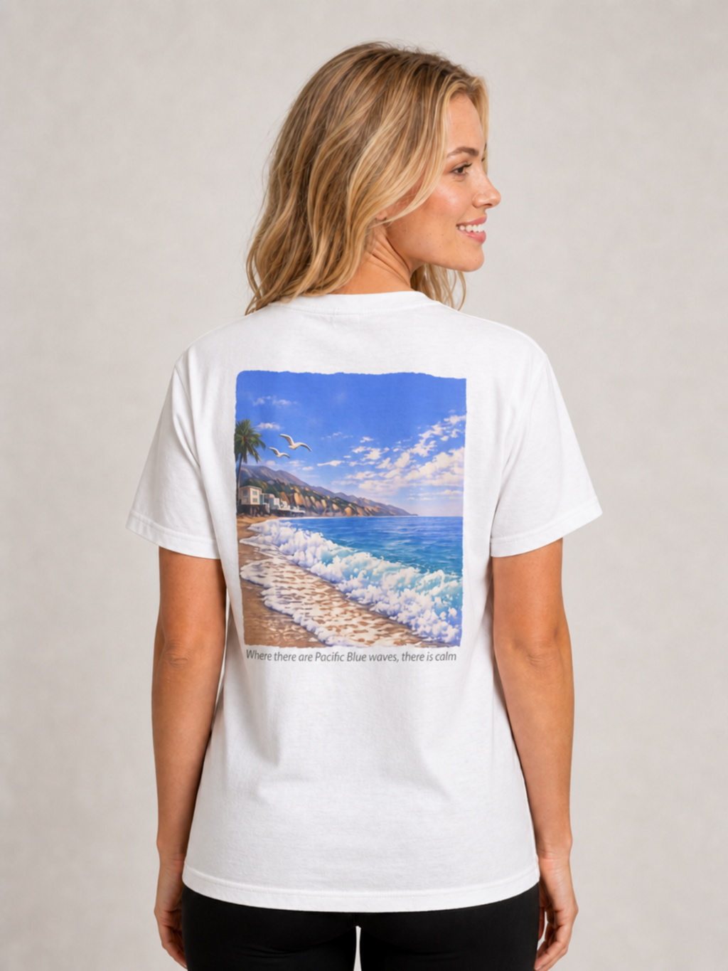 A woman shown from the back wearing a white short-sleeve T-shirt with a large square beach scene graphic on the back. The graphic depicts blue ocean waves, a sandy shore, palm trees, buildings, and a bright blue sky. Below the graphic, the text reads 'Where there are Pacific Blue waves, there is calm'.