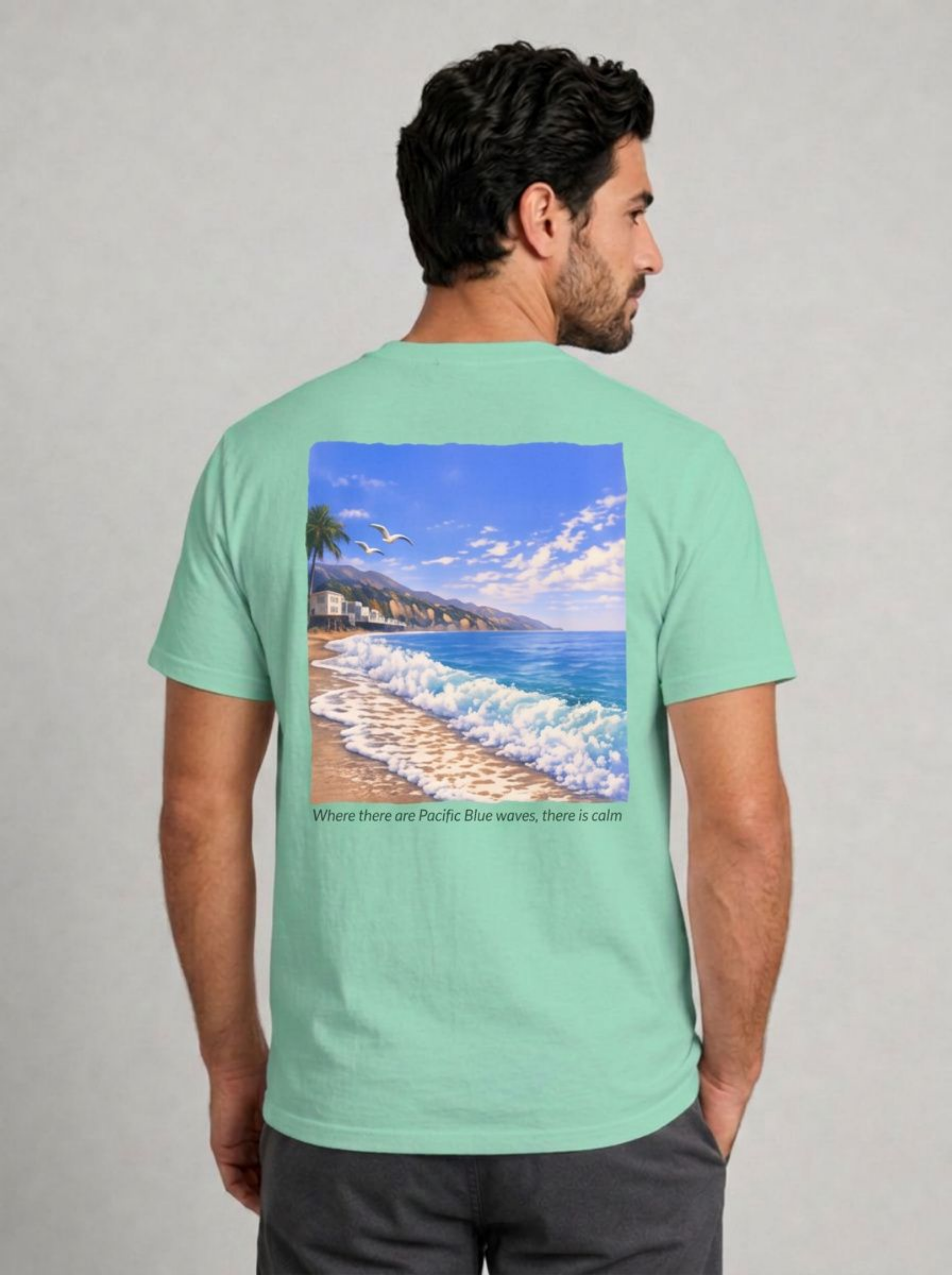 A man wearing a mint green t-shirt, viewed from the back. The shirt features a large square graphic print of a beach scene with blue waves, white surf, and a sandy shore, with the text 'Where there are Pacific Blue waves, there is calm' printed below the graphic.