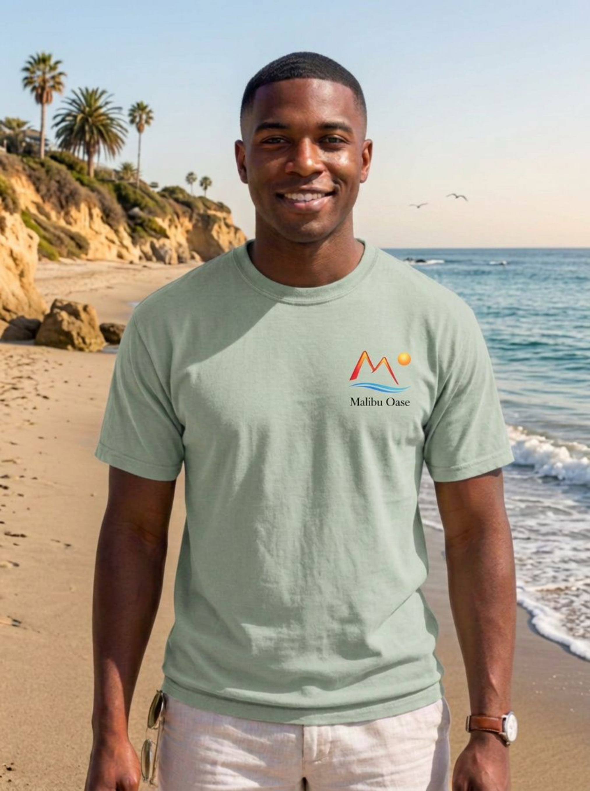 A man standing on a sandy beach wearing a light sage green t-shirt with a small 'Malibu Oasi' logo on the left chest, paired with white pants.