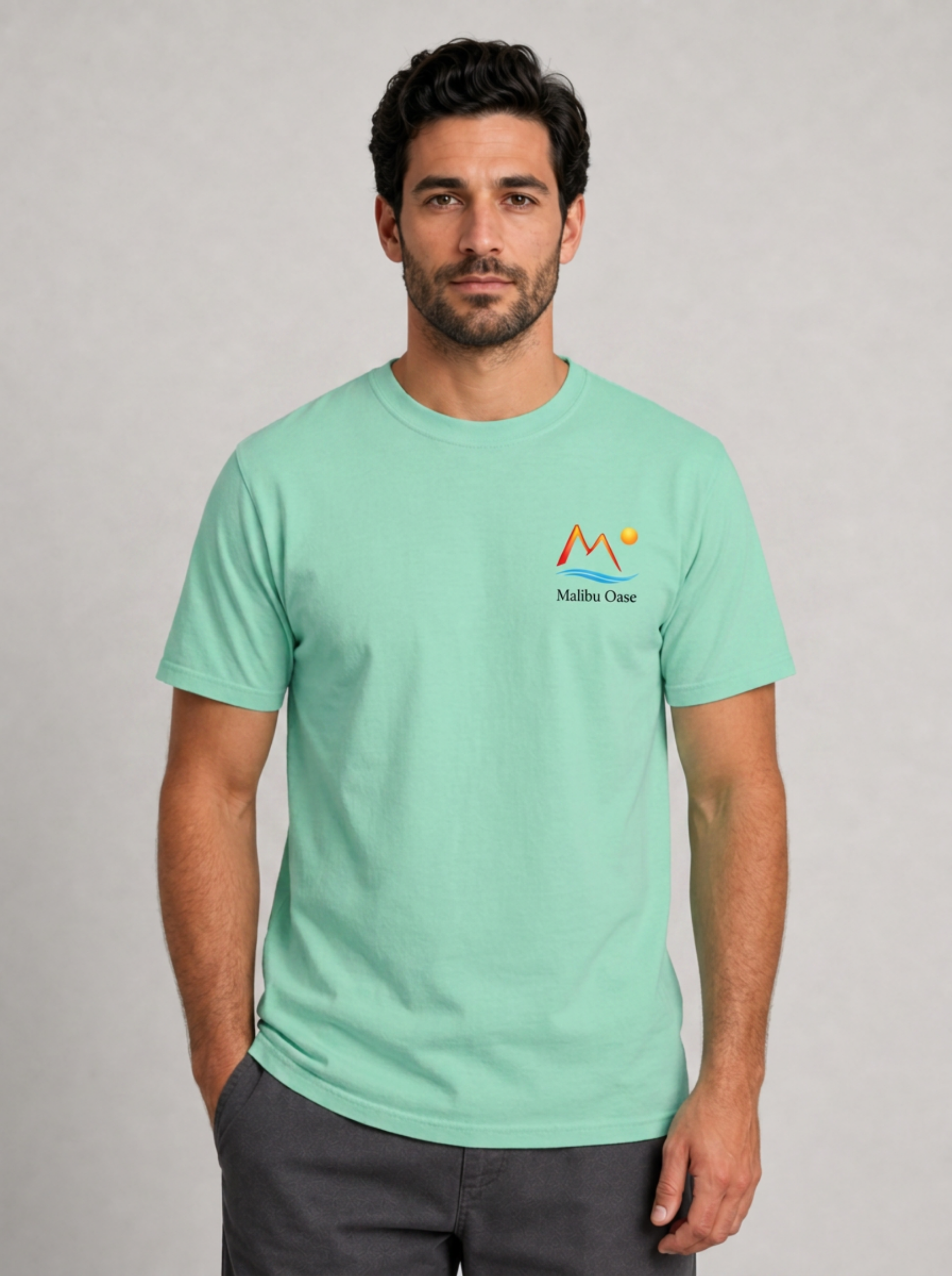 A man wearing a mint green crewneck t-shirt with a small 'Malibu Oase' graphic on the left chest, paired with dark gray pants, standing against a neutral gray background.