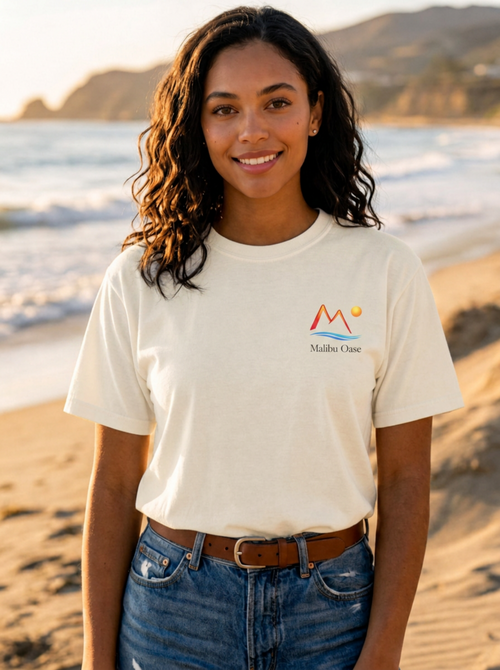 A woman standing on a sandy beach at sunset wearing a cream-colored t-shirt with a red and yellow mountain logo and the text 'Malibu Oasi' on the left chest, paired with blue jeans and a brown belt.