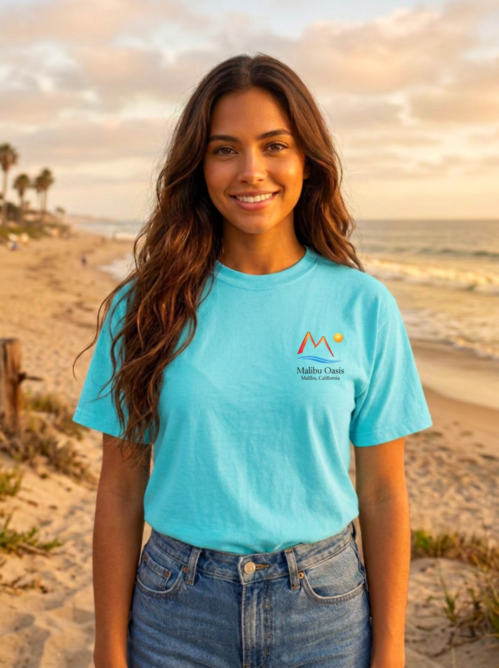 A smiling woman with long brown hair stands on a sandy beach at sunset wearing a bright turquoise short-sleeve T-shirt with a small multicolor mountain logo and the text 'Malibu Oase' on the left chest, paired with blue jeans.