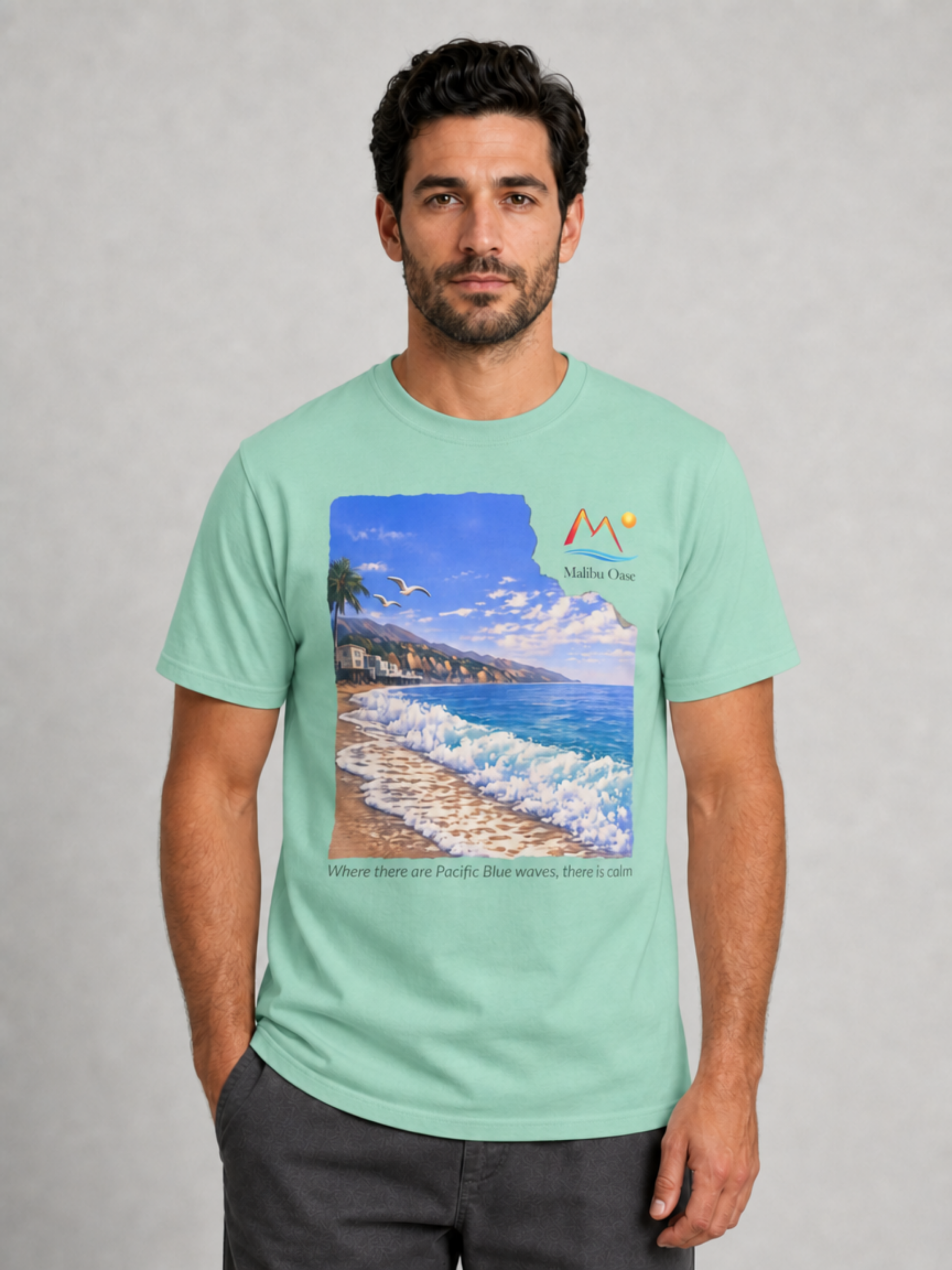 A man wearing a mint green graphic t-shirt with a scenic California beach print and the logo 'Malibu Oase' and under the print there are the words 'Where there are Pacific Blue waves, there is calm'.