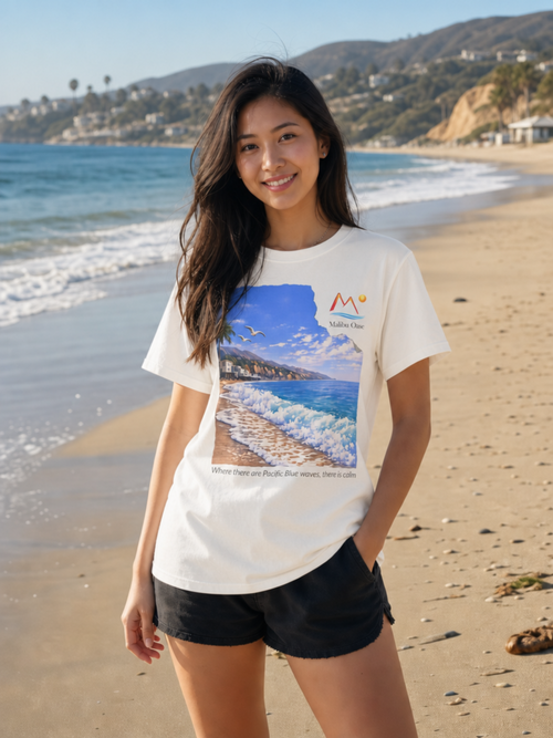 A woman stands on a sandy beach wearing a white graphic T-shirt with a large coastal print and black shorts. The background shows ocean waves, shoreline, hills, and buildings under a clear blue sky.